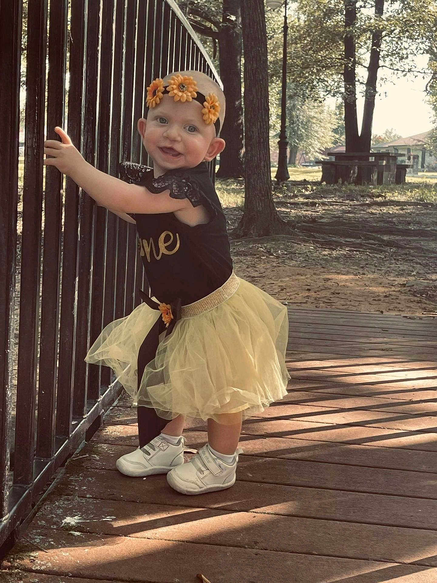 Hazel is registered to the contest to win money with this photo: toddler, child, smiling, outdoor, wooden_deck, metal_railing, yellow_skirt, headband, flowers, white_shoes, sunlight, shadow, trees, grass, park, black_top, happy, person, cute, nature