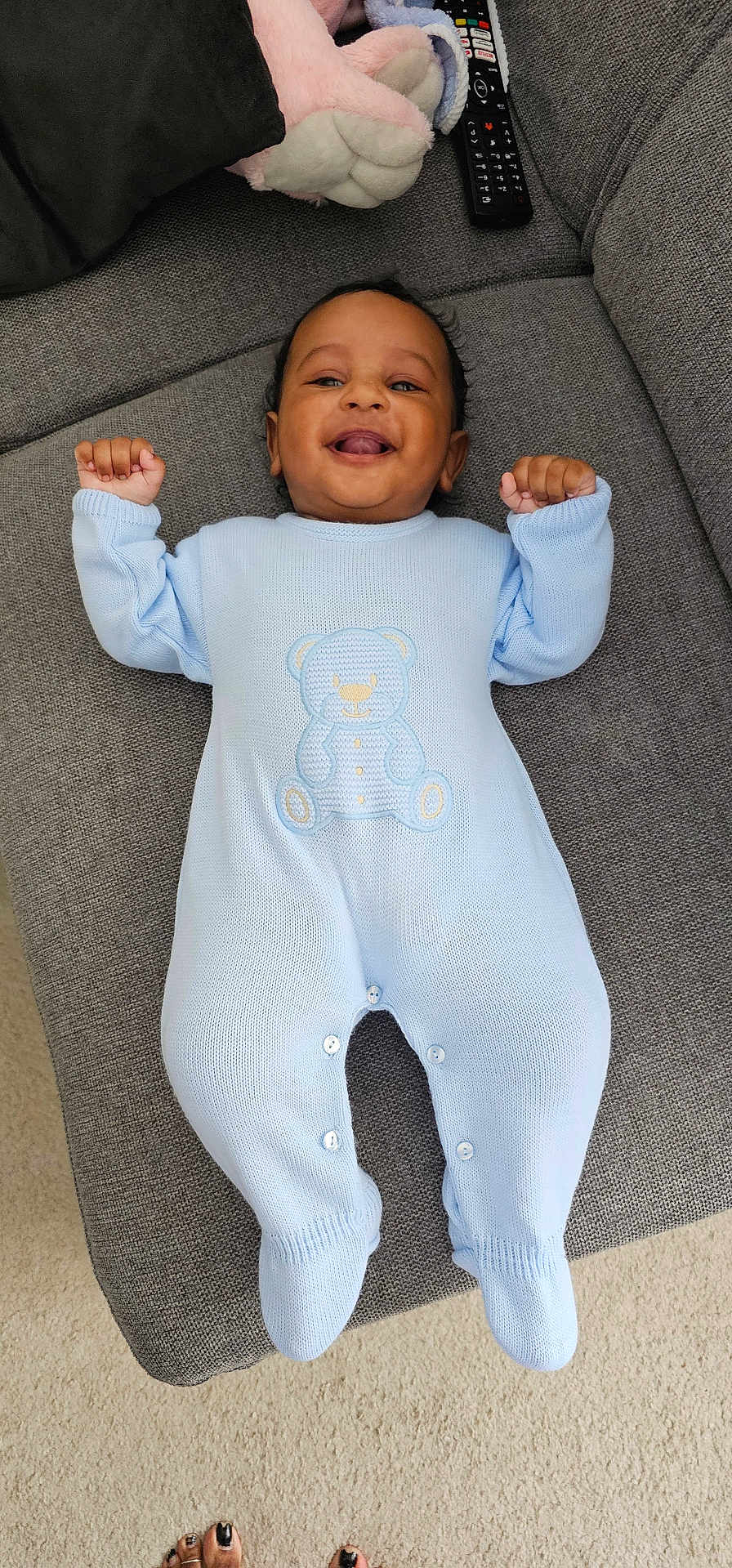 Aarav joined the competition — help win amazing prizes! baby, infant, blue_onesie, teddy_bear_design, smiling, lying_down, couch, gray_couch, plush_toy, remote_control, carpet, happy, raised_arms, indoors, person, footwear, toes, knitted_clothing, cute, portrait