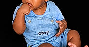 Saage joined the competition — help win amazing prizes! baby, child, infant, overalls, blue_clothing, sitting, black_background, hand_in_mouth, barefoot, cute, portrait, young_child, studio_shot, expression, curious, skin, face, toe, casual, person