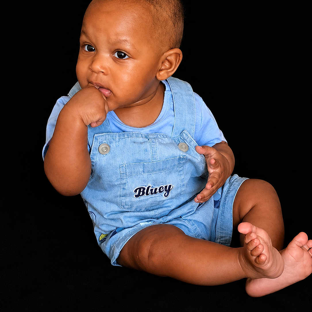 Saage joined the competition — help win amazing prizes! baby, child, infant, overalls, blue_clothing, sitting, black_background, hand_in_mouth, barefoot, cute, portrait, young_child, studio_shot, expression, curious, skin, face, toe, casual, person