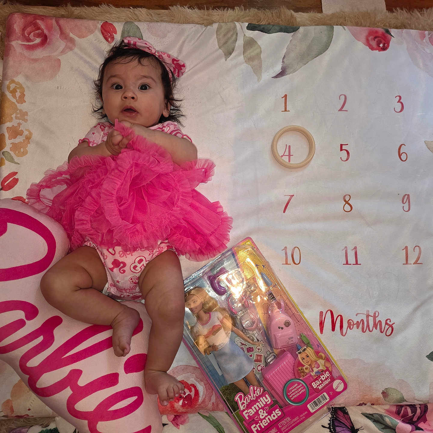 Violet is registered to the contest to win money with this photo: baby, pink_tutu, headband, milestone_blanket, floral_pattern, carpet, barbie_pillow, barbie_doll, toy_packaging, infant, clothing, lying_down, indoors, cute, child, portrait, month_marker, colorful, playtime, decorative