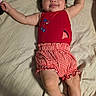 baby, child, smile, lying_down, red_clothing, checkered_shorts, bed, blanket, happy, infant, cute, person, skin, arm, leg, head, face, indoors, soft_lighting, comfortable