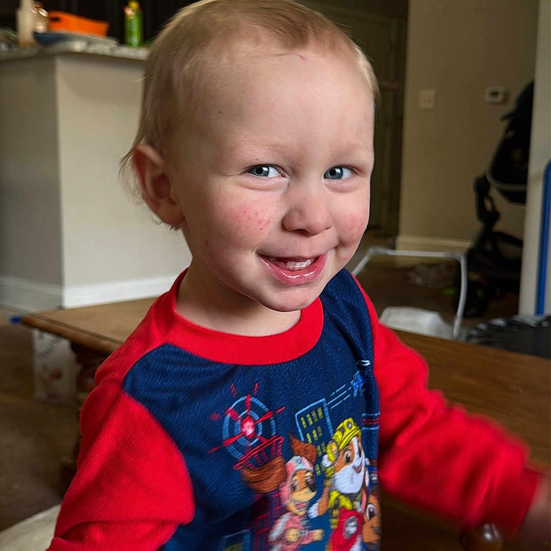 Bradley is registered to the contest to win money with this photo: blue_shirt, cabinet, carpet, child, clothing, face, foot, furniture, happy, indoor, light_fixture, living_room, motion_blur, paw_patrol, person, red_sleeves, smiling, toddler, toy, wooden_table