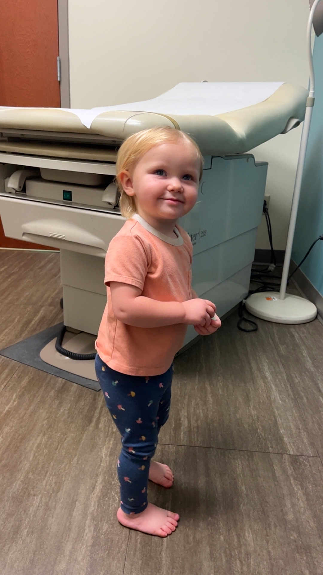 Ella joined the competition — help win amazing prizes! toddler, child, barefoot, standing, medical_clinic, examination_table, floor_lamp, peach_shirt, navy_pants, patterned_clothing, smiling, indoor, flooring, medical_equipment, curly_hair, young_child, waiting, cute, small_person, healthcare