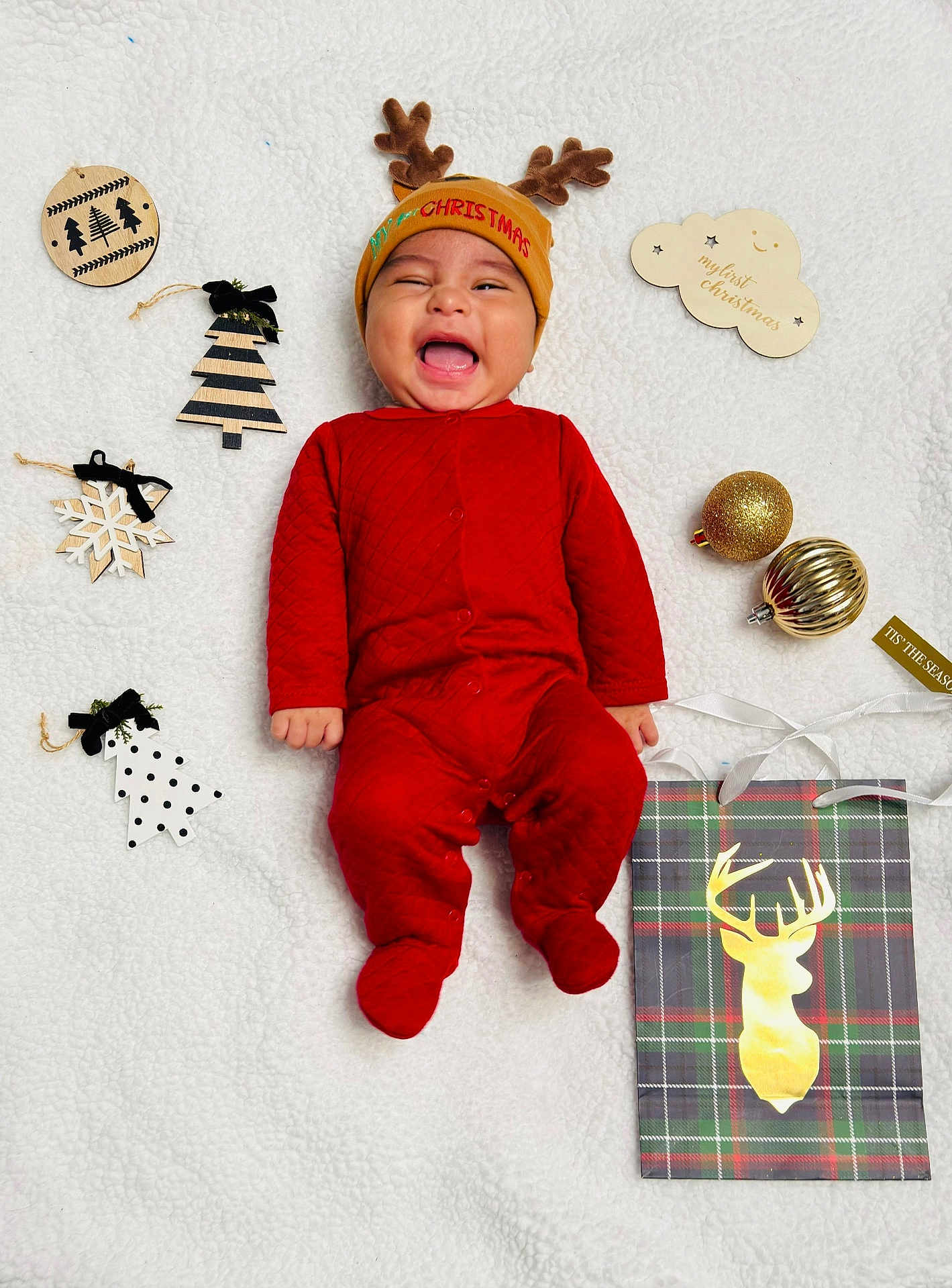 Jake is registered to the contest to win money with this photo: baby, infant, red_onesie, reindeer_hat, christmas_decorations, wooden_ornaments, gold_baubles, white_blanket, smiling, holiday, festive, cute, happy, lying_down, indoors, portrait, seasonal, celebration, child, cozy