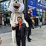 child, boy, mascot, bunny, costume, peace_sign, smile, outdoor, urban, building, sidewalk, jacket, sneakers, person, casual, daylight, happy, background, signage, glasses