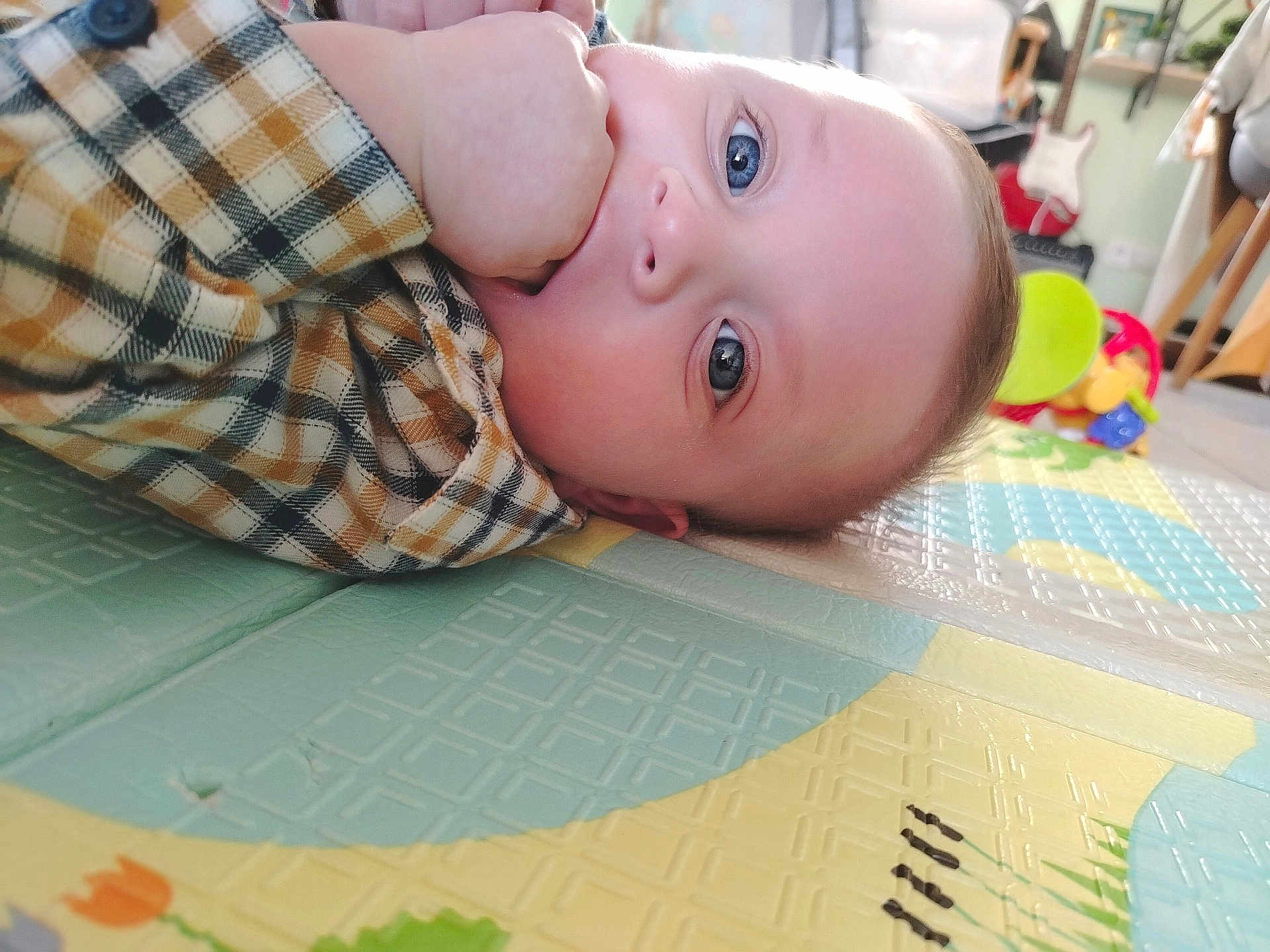 Gaël a rejoint le concours — aidez-le/la à gagner de superbes lots ! baby, child, blue_eyes, hand, plaid_shirt, play_mat, toy, guitar, indoor, floor, face, person, closeup, sitting, cute, infant, home, colorful, curious, young