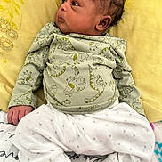 Ryden is registered to the contest to win money with this photo: newborn, baby, infant, child, portrait, face, eyes, hair, lying_down, blanket, yellow_bedding, dinosaur_print, clothing, white_pants, hands, indoor, bed, cozy, cute, photography