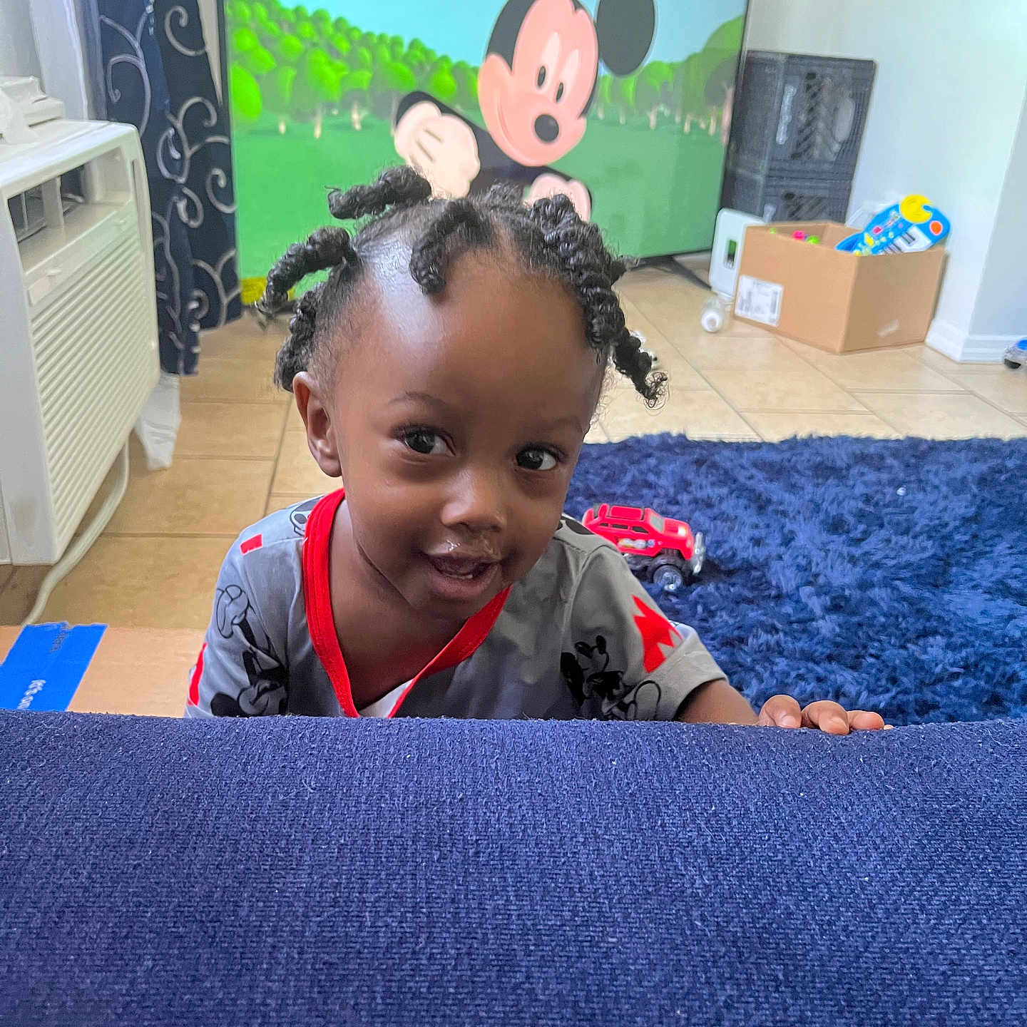 Jiaie is registered to the contest to win money with this photo: air_conditioner, blue_couch, box, carpet, cartoon, child, curly_hair, curtain, floor_tiles, furniture, home, indoor, living_room, mickey_mouse, person, playful, smiling, television, toddler, toy_car