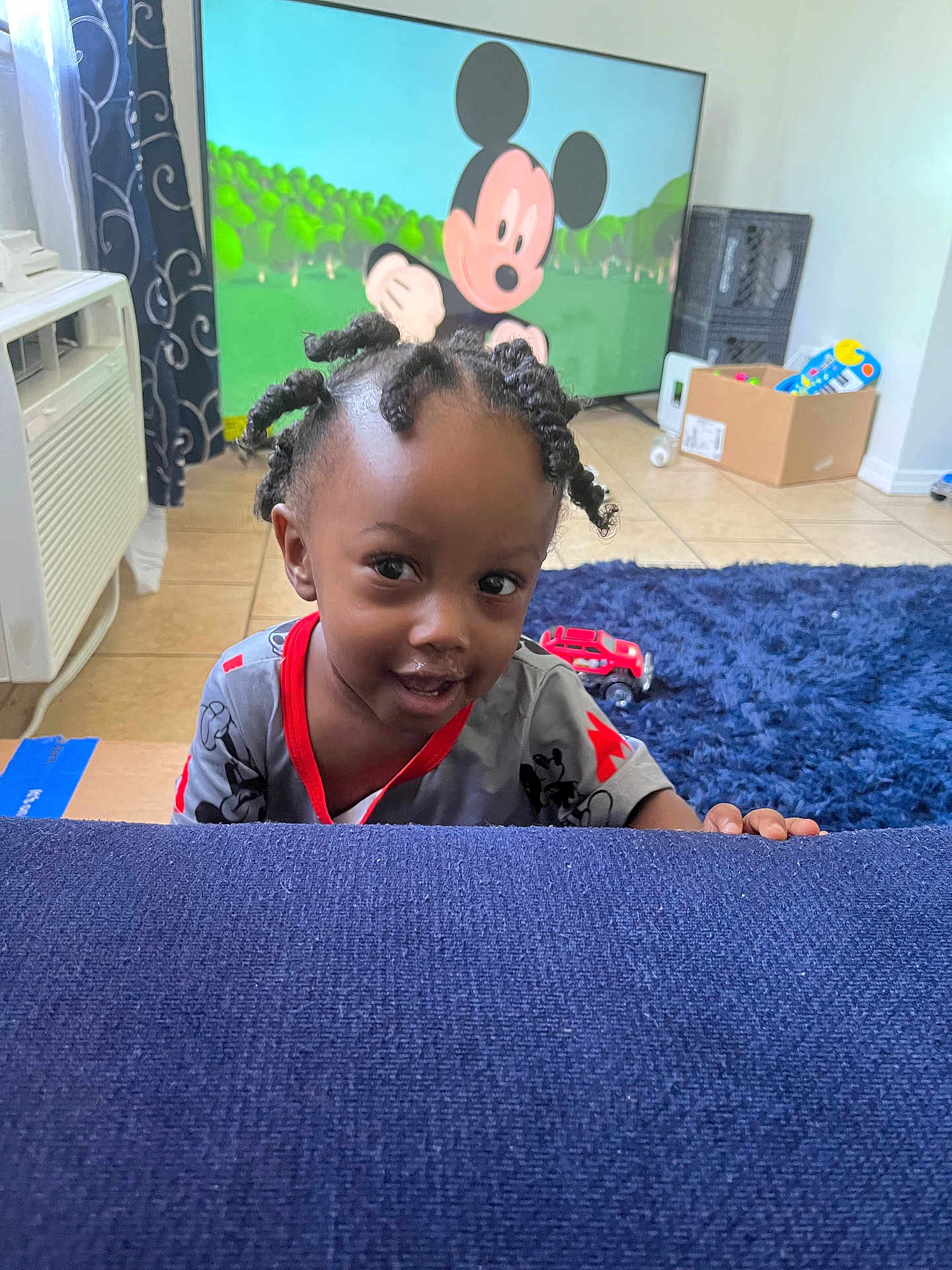 Jiaie is registered to the contest to win money with this photo: air_conditioner, blue_couch, box, carpet, cartoon, child, curly_hair, curtain, floor_tiles, furniture, home, indoor, living_room, mickey_mouse, person, playful, smiling, television, toddler, toy_car
