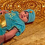 Ishtar is registered to the contest to win money with this photo: baby, infant, blue_clothing, golden_fabric, headwear, lying_down, cute, child, portrait, face, arm, bracelet, bright_colors, texture, soft_lighting, indoors, closeup, expression, decorative, costume
