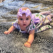 Ishtar is registered to the contest to win money with this photo: baby, infant, purple_headband, floral_dress, blanket, patterned_blanket, indoor, child, cute, lying_down, wide_eyes, open_mouth, curious, soft_lighting, toddler, crawling_position, bracelet, skin, person, portrait