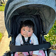 Angie is registered to the contest to win money with this photo: baby, stroller, outdoor, child, curious, pink_pants, white_top, sunlight, sidewalk, greenery, car, seatbelt, evenflo, hand_bracelet, infant, face, portrait, sitting, cute, expression