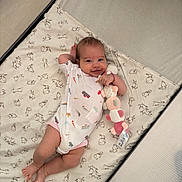 Emmy a rejoint le concours — aidez-le/la à gagner de superbes lots ! adorable, baby, baby_clothes, baby_girl, bedding, child, crib, cute, happy, indoors, infant, lying_down, onesie, patterned_bedding, person, playful, plush_toy, resting, smiling, soft_toy