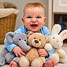 baby, child, smiling, plush_toy, elephant_toy, teddy_bear, bunny_toy, carpet, indoor, basket, blue_clothing, cute, happy, person, toy, furniture, sitting, face, expression, home