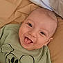 baby, smiling, tongue_out, green_shirt, cartoon_print, lying_down, bed, bedding, happy, infant, cute, child, face, skin, head, eyes, mouth, clothing, indoors, portrait