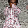 Zuri is registered to the contest to win money with this photo: baby, bed, bedding, child, clothing, cozy, curly_hair, dark_skin, face, floral_onesie, hands, home, indoors, infant, lying_down, patterned_fabric, photography, portrait, sleepwear, wide_eyes