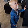 child, toddler, pacifier, blue_overalls, indoor, curious, standing, blonde_hair, long_sleeve_shirt, white_shoes, furniture, wood_floor, home_interior, cabinet, fan, black_couch, pillow, blanket, toy_clip, person