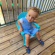 Mathis a rejoint le concours — aidez-le/la à gagner de superbes lots ! toddler, child, blue_chair, curly_hair, wooden_deck, outdoor, sandals, fence, green_fence, romper, casual_clothing, playful, summer, sunlight, plastic_chair, person, young_child, curious, head_tilt, daylight