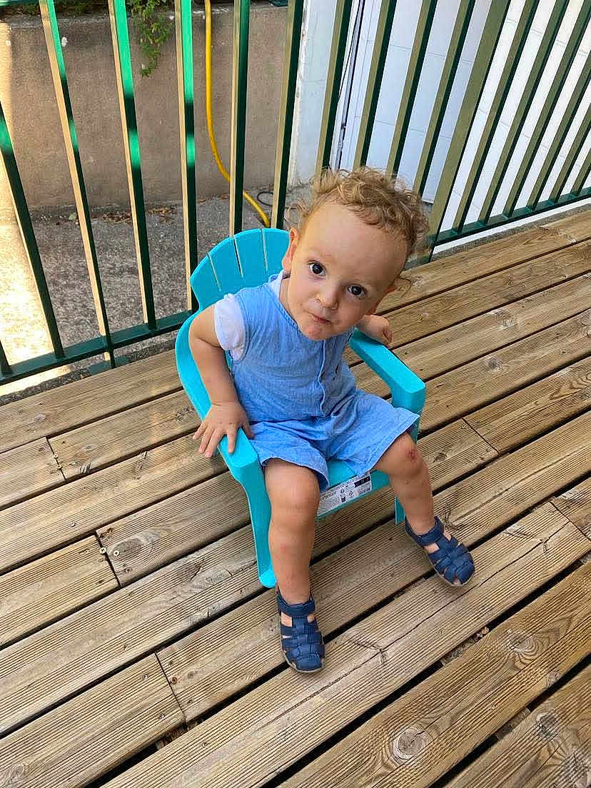 Mathis a rejoint le concours — aidez-le/la à gagner de superbes lots ! toddler, child, blue_chair, curly_hair, wooden_deck, outdoor, sandals, fence, green_fence, romper, casual_clothing, playful, summer, sunlight, plastic_chair, person, young_child, curious, head_tilt, daylight