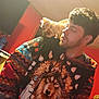 man, cat, hoodie, wolf_design, shoulder, indoor, red_wall, television, facial_hair, curly_hair, looking, pet, animal, casual_clothing, human, small_cat, scruffy_cat, room, warm_lighting, portrait