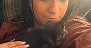 Luna is registered to the contest to win money with this photo: person, kitten, cat, black_cat, hand, cuddle, indoor, closeup, face, long_nails, ring, shirt, plaid_cushion, natural_light, casual_clothing, brown_shirt, pet, cozy, soft_light, relaxation