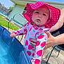 baby, child, swimsuit, hat, pink, polka_dot, strawberry_pattern, pool, water, summer, outdoor, sunny, grass, house, adult_hand, curious, daylight, backyard, person, footwear