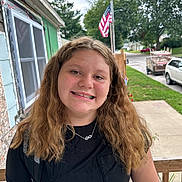 Adison is registered to the contest to win money with this photo: child, girl, smile, portrait, outdoor, porch, american_flag, backpack, casual_clothing, black_shirt, necklace, wavy_hair, suburb, sidewalk, trees, car, residential, daytime, happy, person