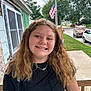 child, girl, smile, portrait, outdoor, porch, american_flag, backpack, casual_clothing, black_shirt, necklace, wavy_hair, suburb, sidewalk, trees, car, residential, daytime, happy, person