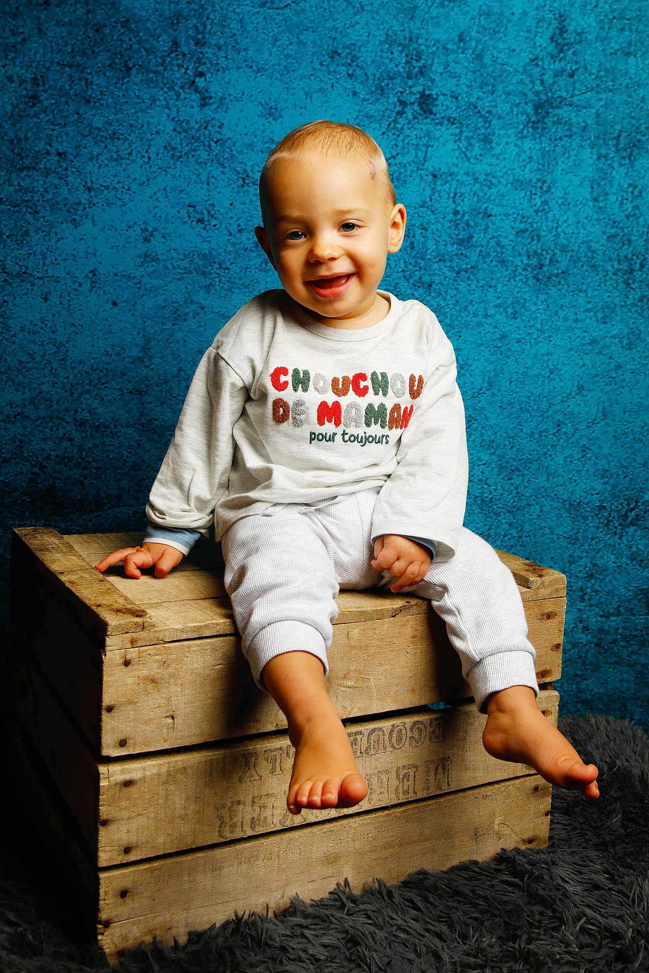 Nino a rejoint le concours — aidez-le/la à gagner de superbes lots ! toddler, child, smiling, barefoot, wooden_crate, sweater, clothing, blue_background, indoor, portrait, happy, cute, studio_lighting, floor_rug, baby, sitting, person, face, head, casual