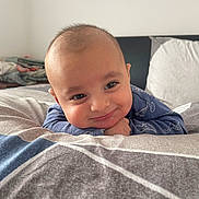 Ibrahim is registered to the contest to win money with this photo: baby, child, face, smile, blue_clothing, bed, pillow, blanket, indoor, portrait, cute, dinosaur_print, head, resting, happy, person, young_child, infant, cozy, room
