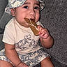 baby, child, dinosaur, hat, bucket_hat, cookie, gingerbread, couch, clothing, pattern, sitting, cute, happy, indoor, portrait, hand, foot, skin, face, chewing