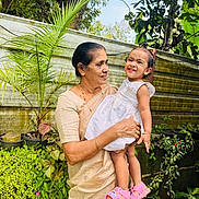Juliet Mariam is registered to the contest to win money with this photo: elderly_woman, toddler, child, woman, smile, white_dress, pink_sandals, garden, plants, greenery, potted_plants, outdoor, nature, fence, portrait, holding, happy, family, person, casual_clothing