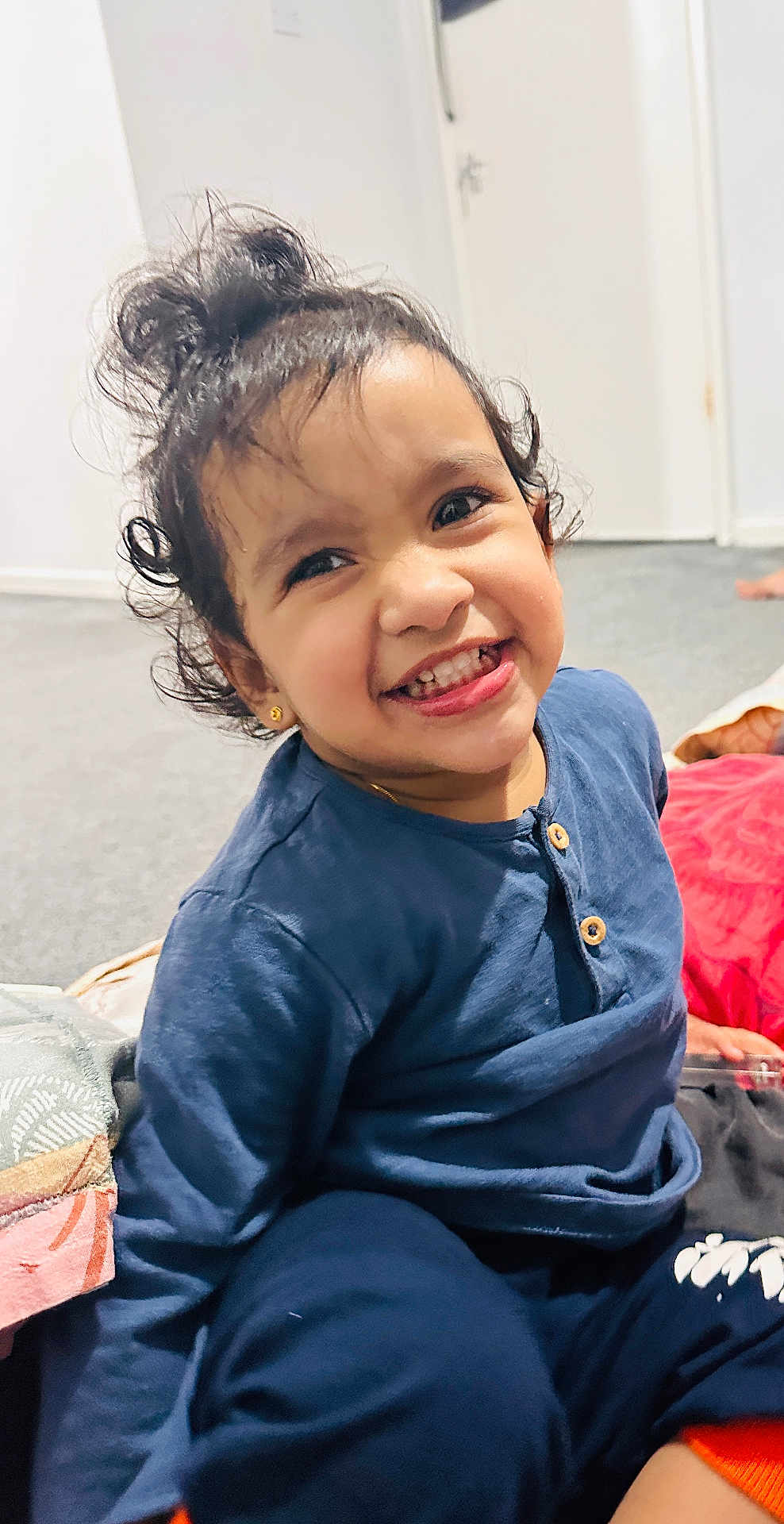 Juliet Mariam is registered to the contest to win money with this photo: child, toddler, smiling, curly_hair, blue_clothing, indoor, carpet, blanket, earrings, happy, person, cute, portrait, sitting, young_child, playful, casual, home, expression, fun