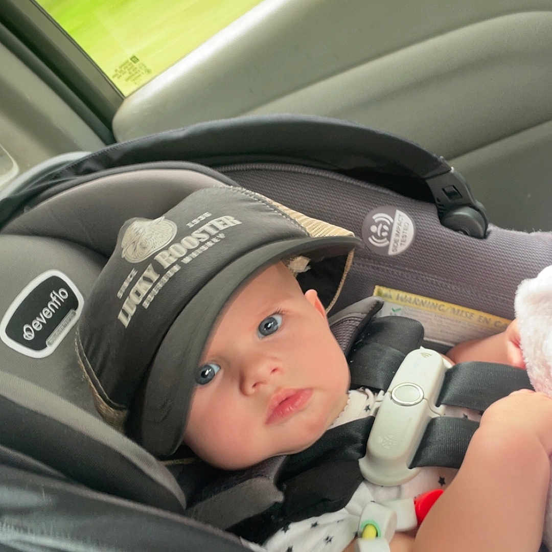 Shyanne is registered to the contest to win money with this photo: baby, car_seat, cap, blue_eyes, toy, seatbelt, infant, child, car_interior, window, curious, sitting, safety, portrait, cute, young_child, clothing, daylight, travel, car