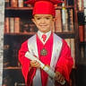 child, graduation, cap, gown, diploma, smiling, portrait, bookshelf, red_clothing, medal, tie, celebration, education, young, person, formal, indoor, photograph, background, marble_surface