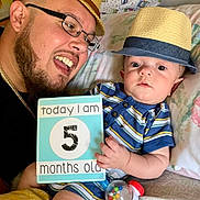 Izaiah joined the competition — help win amazing prizes! baby, adult, hat, glasses, smiling, striped_clothing, sign, toy, bed, pillow, person, infant, happy, child, indoor, portrait, feeding_bottle_toy, closeup, family, casual
