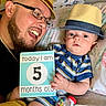 baby, adult, hat, glasses, smiling, striped_clothing, sign, toy, bed, pillow, person, infant, happy, child, indoor, portrait, feeding_bottle_toy, closeup, family, casual