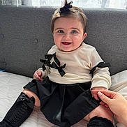 Vivienne is registered to the contest to win money with this photo: baby, child, smiling, sitting, bed, hand_holding, black_bow, cream_sweater, black_skirt, black_socks, curtains, gray_couch, indoor, person, cute, happy, portrait, clothing, infant, holding_hand