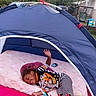 child, toddler, sleeping, smiling, tent, play_tent, outdoor, backyard, mattress, pillow, pajamas, striped_pants, cartoon_clothing, toy, bottle, hand_raised, nap, camping, residential_area, portrait