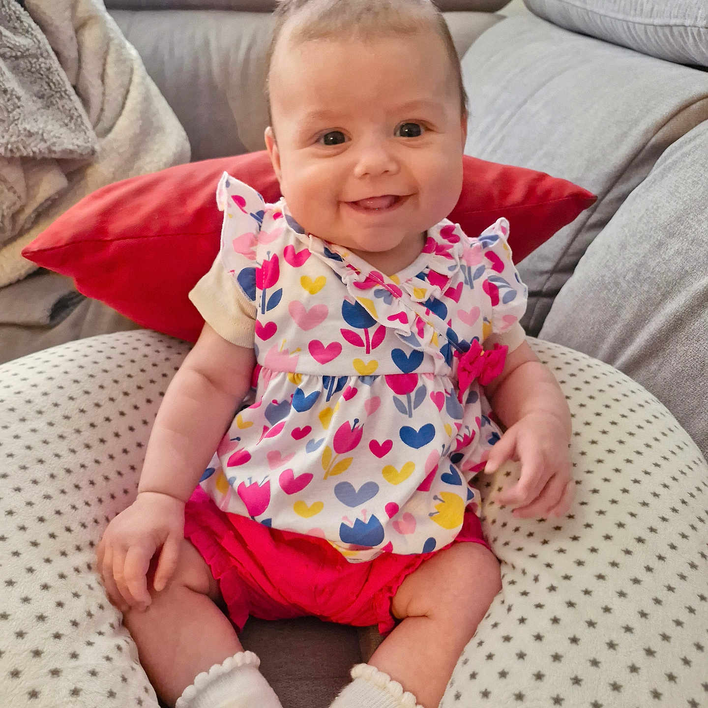 Tessa participe au concours pour gagner de l'argent avec cette photo : baby, blanket, child, clothing, couch, cozy, cute, face, flower_pattern, happy, heart_pattern, indoor, infant, person, pillow, red_shorts, sitting, smiling, sock, white_socks