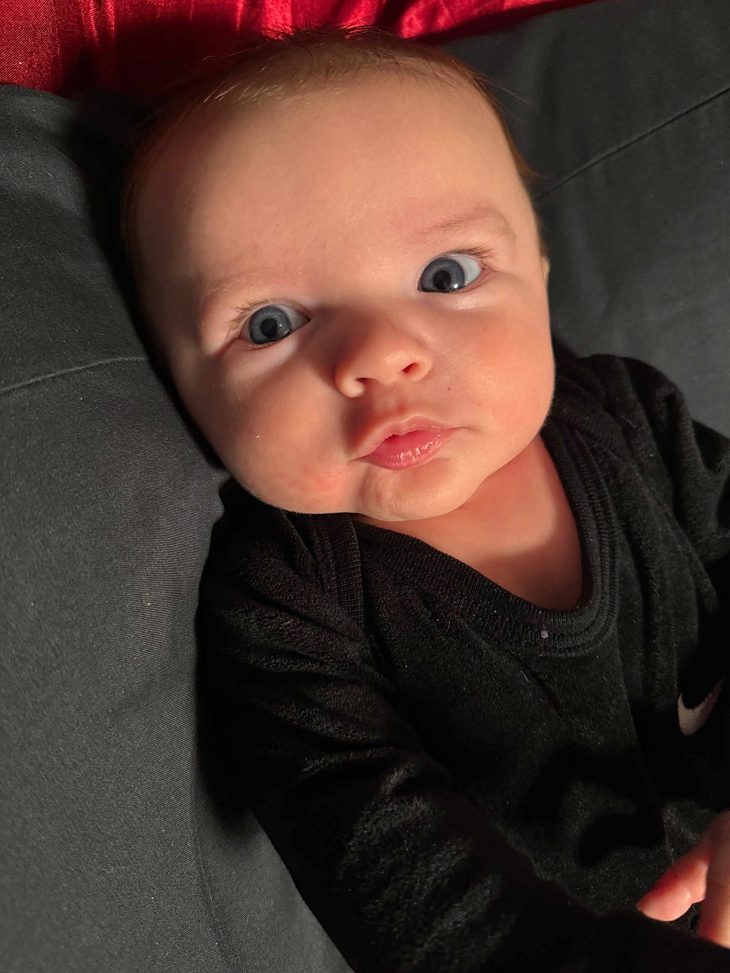 Colton is registered to the contest to win money with this photo: baby, infant, face, eyes, black_clothing, pout, close_up, portrait, child, young, cute, skin, expression, lying_down, indoor, fabric, red_background, soft_light, person, newborn