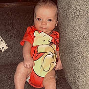 Sonny is registered to the contest to win money with this photo: baby, infant, couch, onesie, orange_clothing, cartoon_character, bear, indoor, furniture, statue, red_cup, relaxing, child, portrait, sitting, cute, person, home, soft_lighting, casual