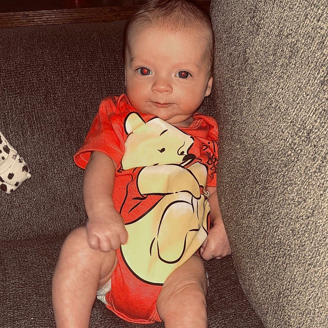 Sonny is registered to the contest to win money with this photo: baby, infant, couch, onesie, orange_clothing, cartoon_character, bear, indoor, furniture, statue, red_cup, relaxing, child, portrait, sitting, cute, person, home, soft_lighting, casual