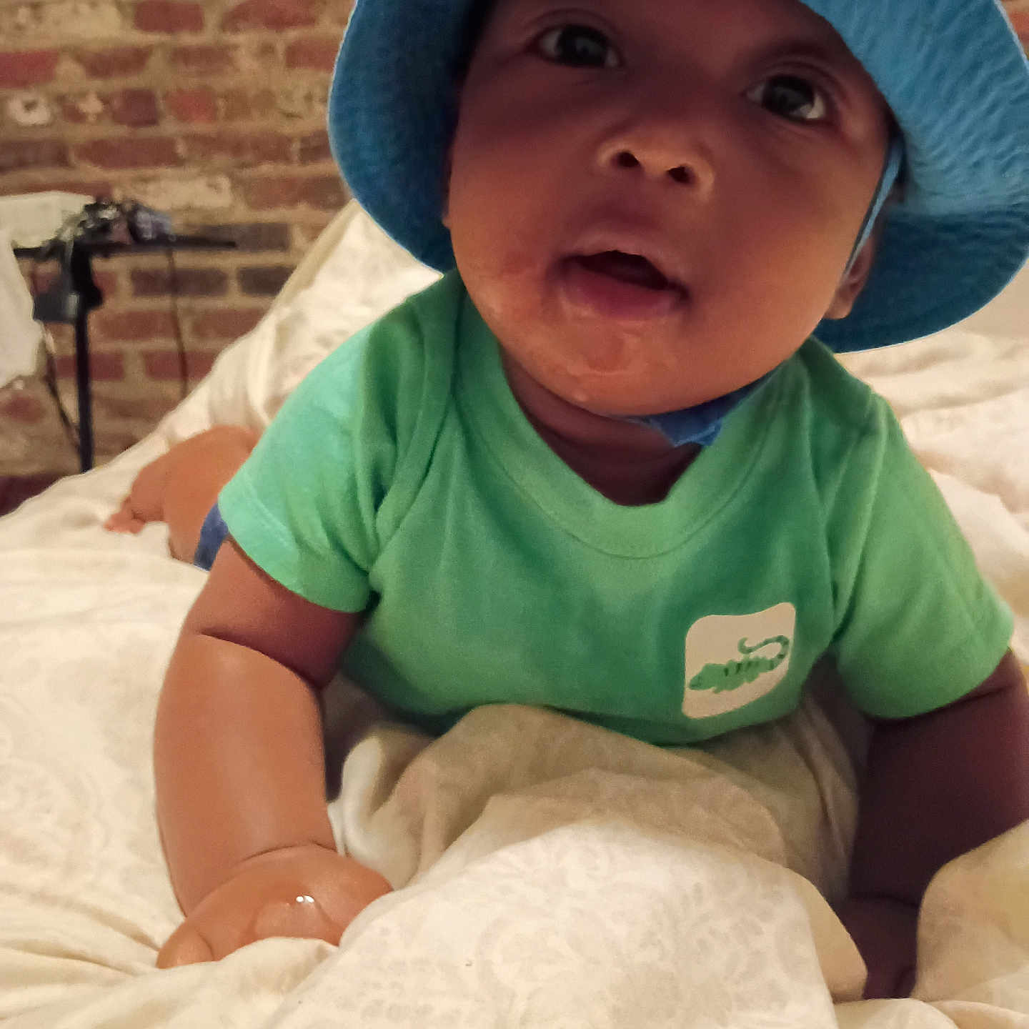 Cameron is registered to the contest to win money with this photo: baby, bed, blanket, blue_hat, brick_wall, child, curious, cute, expression, face, green_shirt, headwear, indoor, infant, lying_down, person, portrait, skin, toddler, young