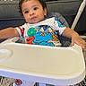 Adonis is registered to the contest to win money with this photo: baby, carpet, child, colorful_clothing, curious, curly_hair, cushion, cute, furniture, high_chair, indoor, living_room, pacifier, pajamas, person, plastic_tray, seated, sesame_street, sofa, toddler