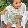 toddler, child, smiling, curly_hair, white_shirt, green_overalls, green_socks, white_chair, outdoor, plants, leaves, ground, soil, natural_light, happy, sitting, cute, portrait, baby, nature
