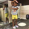 Queani is registered to the contest to win money with this photo: bathroom, bright_colors, casual_clothing, child, countertop, crocs, grey_tshirt, indoor, mirror, person, playful_pose, reflection, sink, smiling, soap_dispenser, standing, tile_floor, tiled_wall, yellow_shorts, young_child