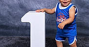 Kyrie is registered to the contest to win money with this photo: toddler, child, smiling, blue_shorts, blue_shirt, white_sneakers, headband, number_one, standing, studio_background, carpet, portrait, happy, cute, indoor, person, baby, sporty, photography, pose