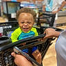 toddler, child, shopping_cart, smile, curly_hair, supermarket, person, hand, smartphone, casual_clothing, indoor, grocery_store, happy, portrait, retail, baby, grip, bright_clothing, blurred_background, candid
